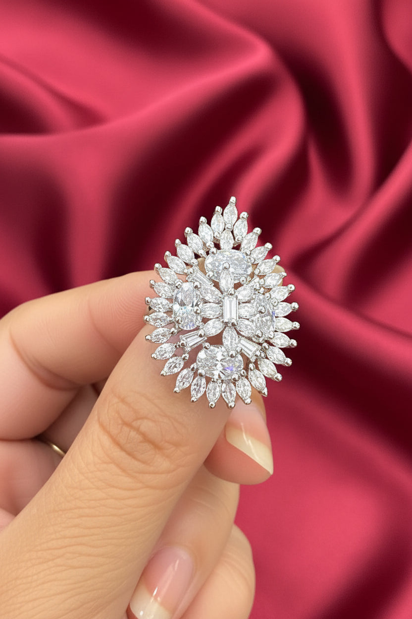 A close-up of a hand holding a large, ornate teardrop-shaped silver ring set with numerous clear gemstones, photographed against a dark brown background.