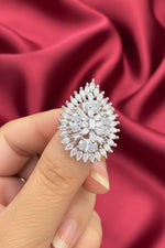 A close-up of a hand holding a large, ornate teardrop-shaped silver ring set with numerous clear gemstones, photographed against a dark brown background.