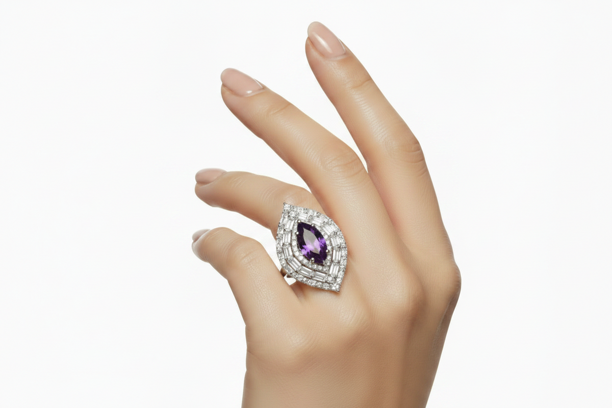 A hand wearing a silver-toned ring featuring a teardrop-shaped purple amethyst center stone surrounded by a halo of small clear stones, set against a plain white background.