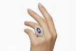A hand wearing a silver-toned ring featuring a teardrop-shaped purple amethyst center stone surrounded by a halo of small clear stones, set against a plain white background.