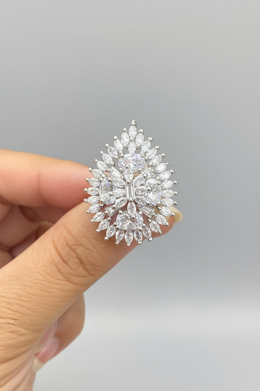 A close-up of a hand holding a silver-toned ring featuring a large, intricate floral cluster of clear marquise and round-cut gemstones.