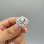 A close-up of a hand holding a silver-toned ring with a large, tiered cluster of clear stones. The ring features a prominent rectangular centre stone and is shown against a blurred background of a grey herringbone floor and a dark brown surface.