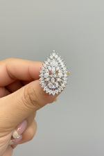 A close-up of a hand holding a silver ring with a large, leaf-like cluster of clear gemstones. The ring is shown against a dark brown background.