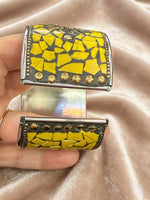 A close-up of a hand holding a wide cuff bracelet with a yellow mosaic tile pattern and gold-toned metal details, set against a draped champagne satin fabric background.