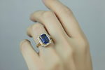 A close-up of a hand wearing a gold plated ring featuring a rectangular sapphire blue-cut centre stone surrounded by a halo of small clear stones, set against a plain light grey background.