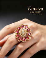 A close-up of a hand wearing a large floral-shaped ring by Famaza Couture. The ring features pink gemstones in teardrop shapes around a central clear stone, set in yellow gold with small clear accent stones.