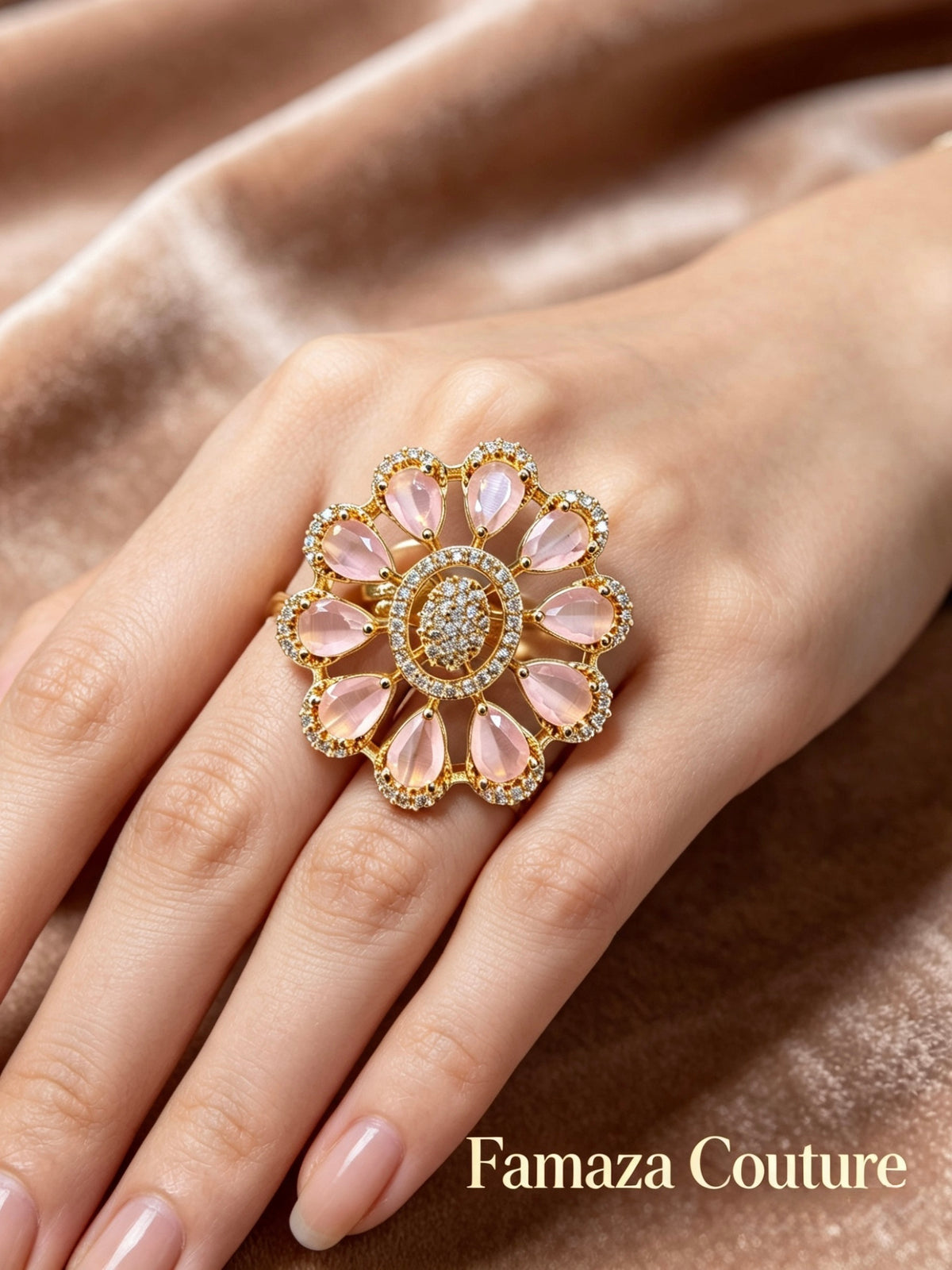 A close-up of a hand wearing a large floral ring by Famaza Couture. The ring features a gold-tone setting with light pink teardrop-shaped stones and a central cluster of small clear stones, set against a soft brown satin background.