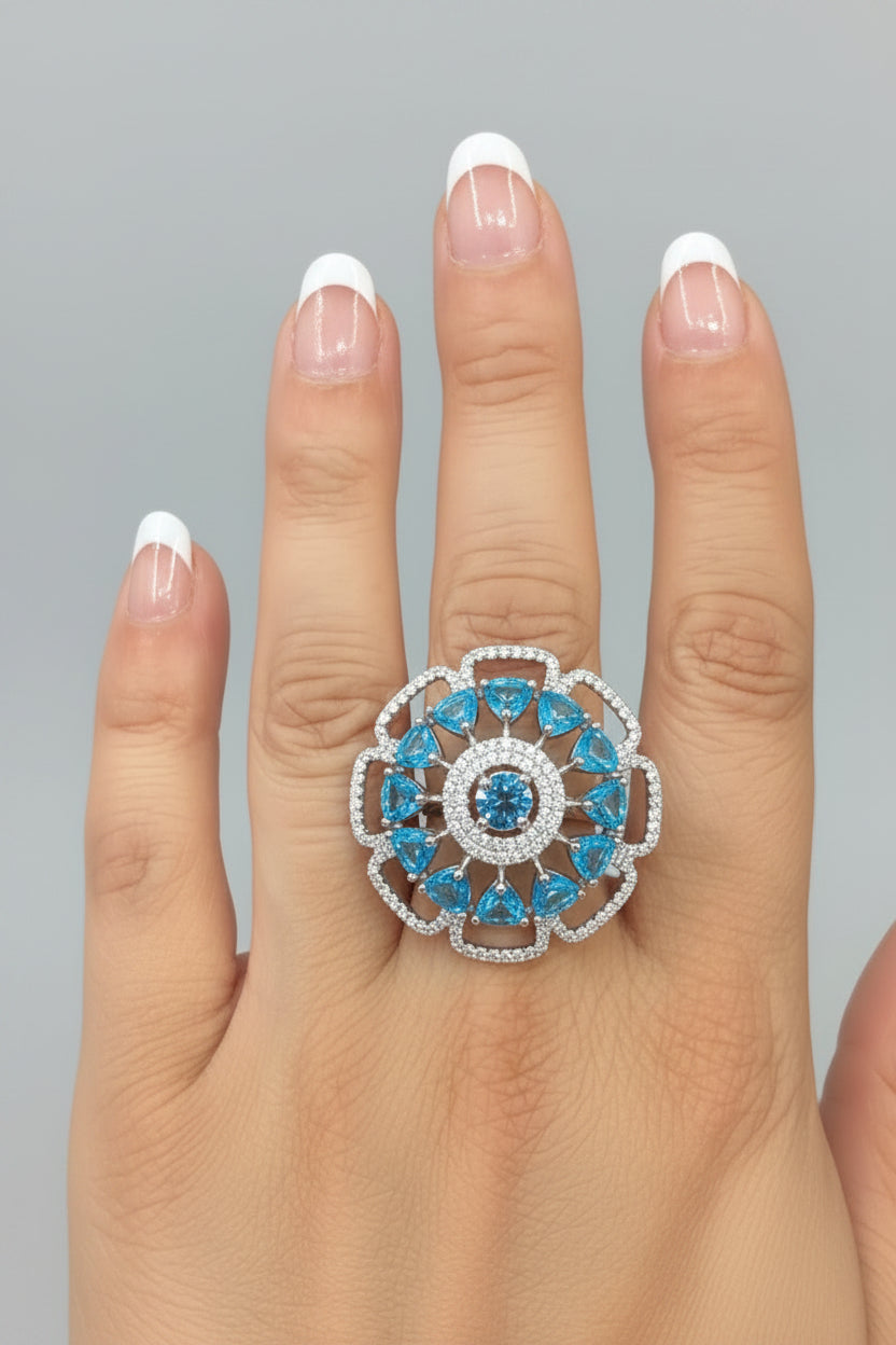 A close-up of a hand wearing a large floral ring with bright blue gemstones and silver-toned metal, set against a black background.