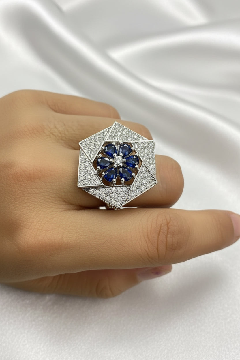 A close-up of a hand wearing a large geometric ring. The ring features a silver-toned hexagonal setting with a central cluster of deep blue round-cut stones surrounded by pavé-set clear stones, with a smaller matching ring on the adjacent finger.