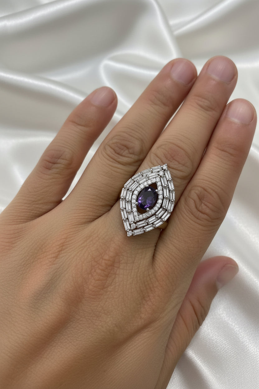 A close-up of a hand wearing a large teardrop-shaped ring with a deep purple oval centre stone and surrounding silver-toned pavé detailing.