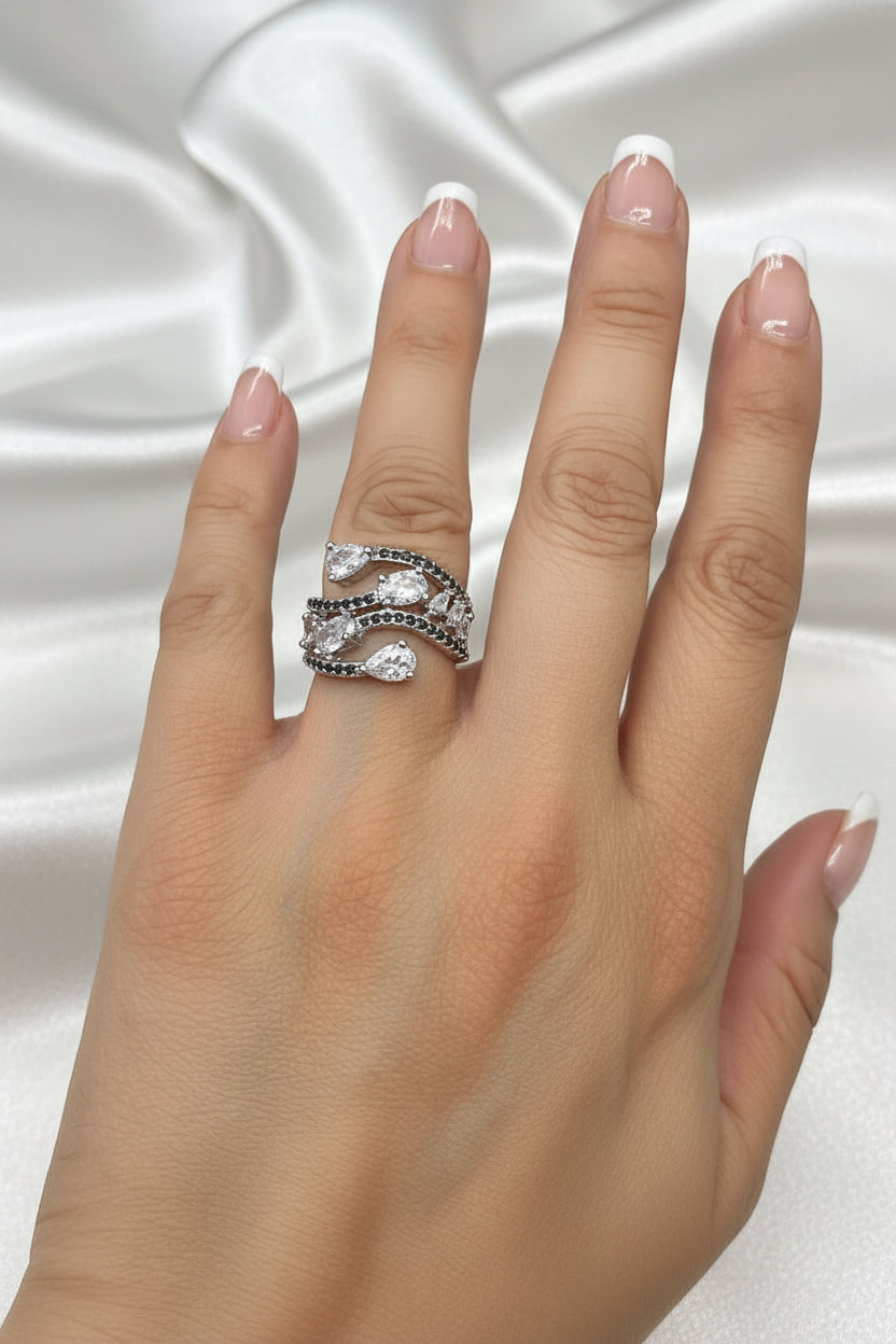 A close-up of a hand wearing a silver-toned wrap ring with clear teardrop-shaped stones and small dark pavé accents, set against a black background.