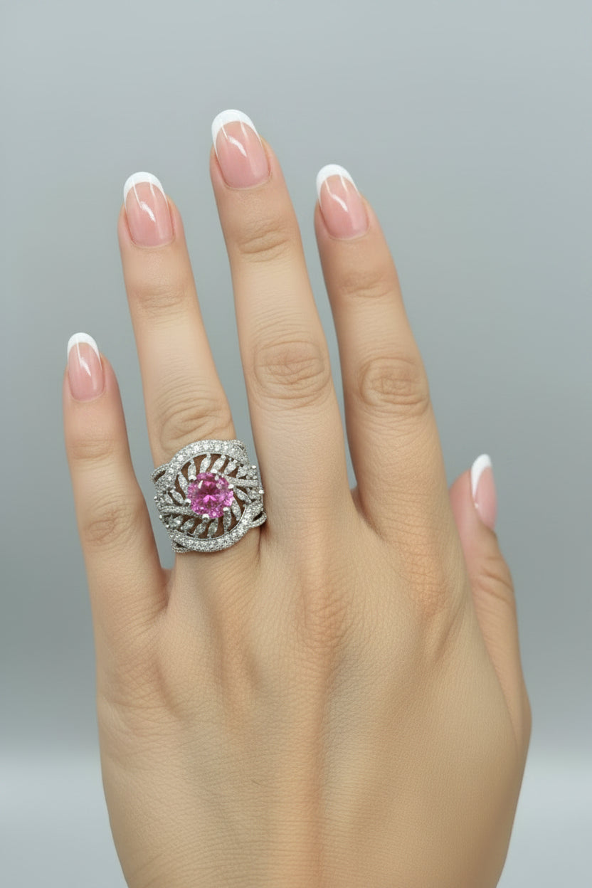 A close-up of a hand wearing a wide silver ring with a central round pink gemstone and surrounding pavé-set silver details, shown against a black background.