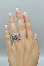 A close-up of a hand wearing a wide silver ring with a central round pink gemstone and surrounding pavé-set silver details, shown against a black background.