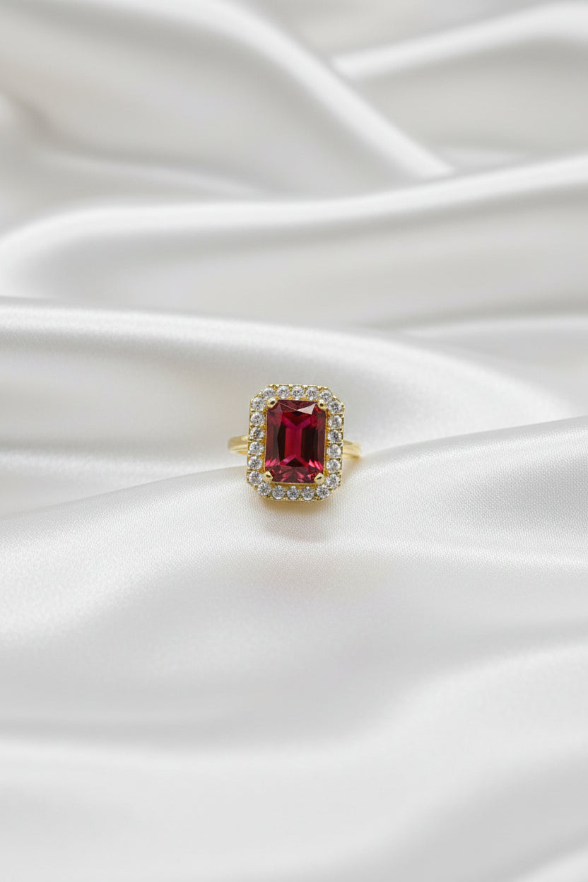 A close-up of a hand wearing a yellow gold ring with a large rectangular red gemstone surrounded by a halo of small clear stones, set against a dark blue textured background.