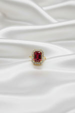 A close-up of a hand wearing a yellow gold ring with a large rectangular red gemstone surrounded by a halo of small clear stones, set against a dark blue textured background.