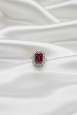 A silver ring featuring a rectangular emerald-cut red gemstone surrounded by a halo of small clear stones, resting on draped white satin fabric.