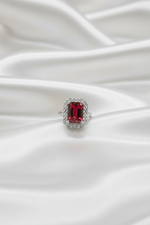 A silver ring featuring a rectangular emerald-cut red gemstone surrounded by a halo of small clear stones, resting on draped white satin fabric.