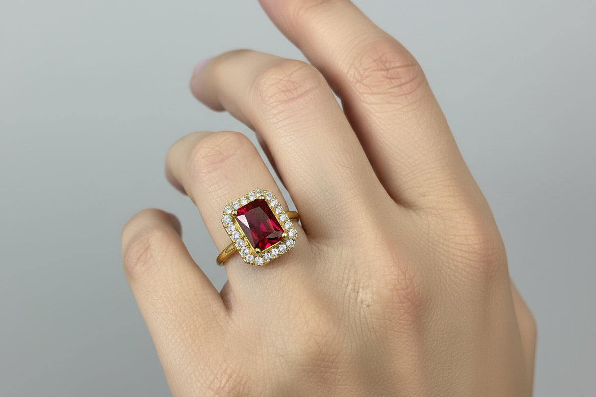 A close-up of a hand wearing a yellow gold ring with a rectangular red gemstone surrounded by a halo of small clear stones, set against a dark blue background.
