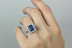 A close-up of a hand wearing a yellow gold ring featuring a rectangular emerald-cut blue sapphire center stone surrounded by a halo of small clear stones, set against a plain light gray background.