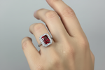 A close-up of a hand wearing a silver-toned ring with a rectangular red gemstone surrounded by a halo of small clear stones, set against a plain light gray background.