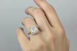 A yellow gold ring with a large emerald-cut center stone and a halo of small round stones, worn on a hand against a soft gray background.