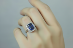 A close-up of a hand wearing a yellow silver plated ring with a rectangular blue gemstone surrounded by a halo of small clear stones, set against a soft grey background.