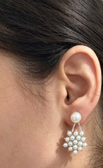 A close-up of a person's ear wearing a dangling pearl earring with a silver-toned floral cluster drop, against a light indoor background.