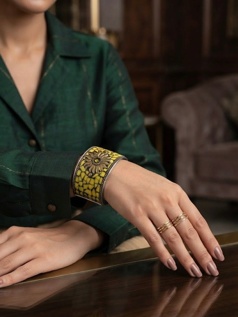 A close-up of a person's hand resting on a polished wooden table, wearing a wide silver cuff bracelet with a yellow and black floral mosaic pattern and gold rings.