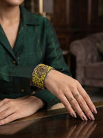 A close-up of a person's hand resting on a polished wooden table, wearing a wide silver cuff bracelet with a yellow and black floral mosaic pattern and gold rings.