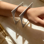 A close-up of a person's wrist wearing a pair of large silver-toned triangular cuff bracelets with ornate engraved detailing.