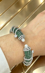 A close-up of a person's wrist wearing two matching cuff-style bracelets. The bracelets feature rows of deep green stones, silver-toned pavé detailing, and a single white pearl at the front of each.