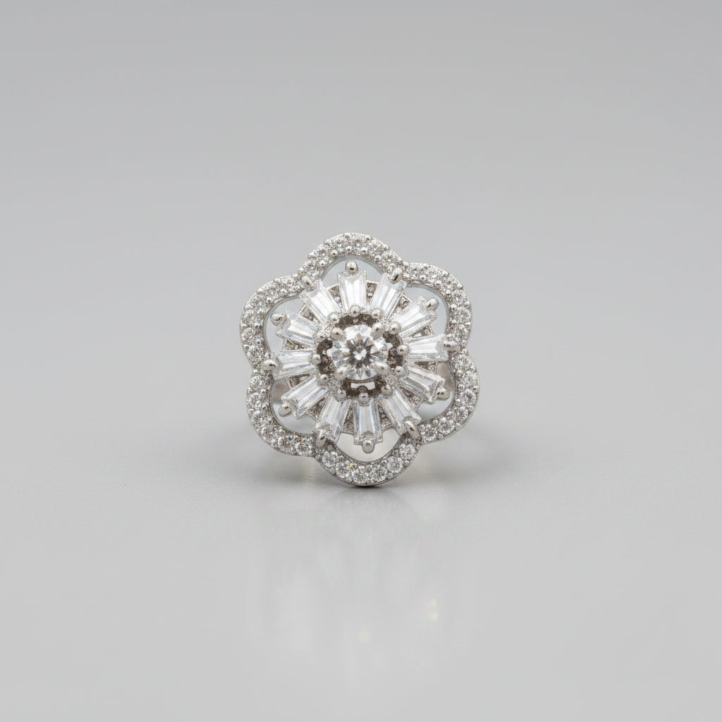A close-up of a silver-toned flower-shaped ring with a central round diamond and surrounding baguette and round diamonds, held against a dark display cone with fingers visible in the background.