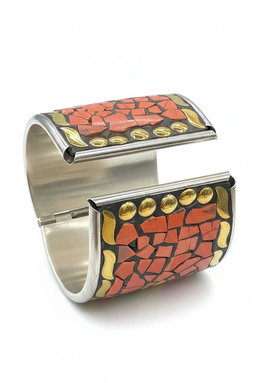 A close-up of a wide cuff bracelet being held between fingers. The bracelet features a mosaic pattern of orange-red and gold pieces set in silver-tone metal, with a large silver clasp visible in the centre.