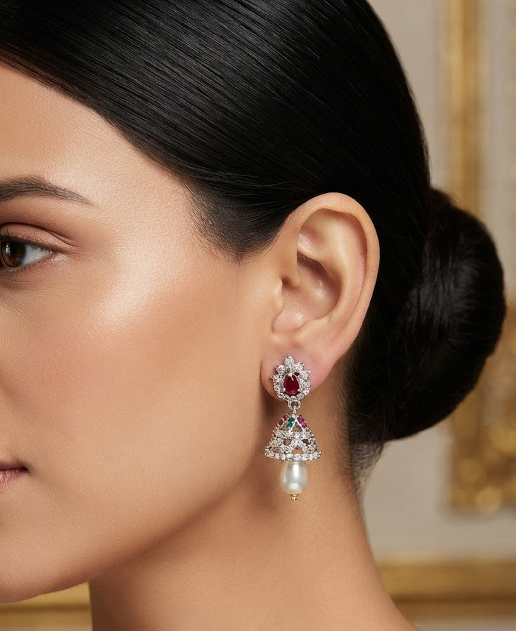 A close-up of a woman wearing a dangling chandelier earring with a red ruby stone, clear pavé setting, and a white pearl drop, shown against a softly blurred interior background.