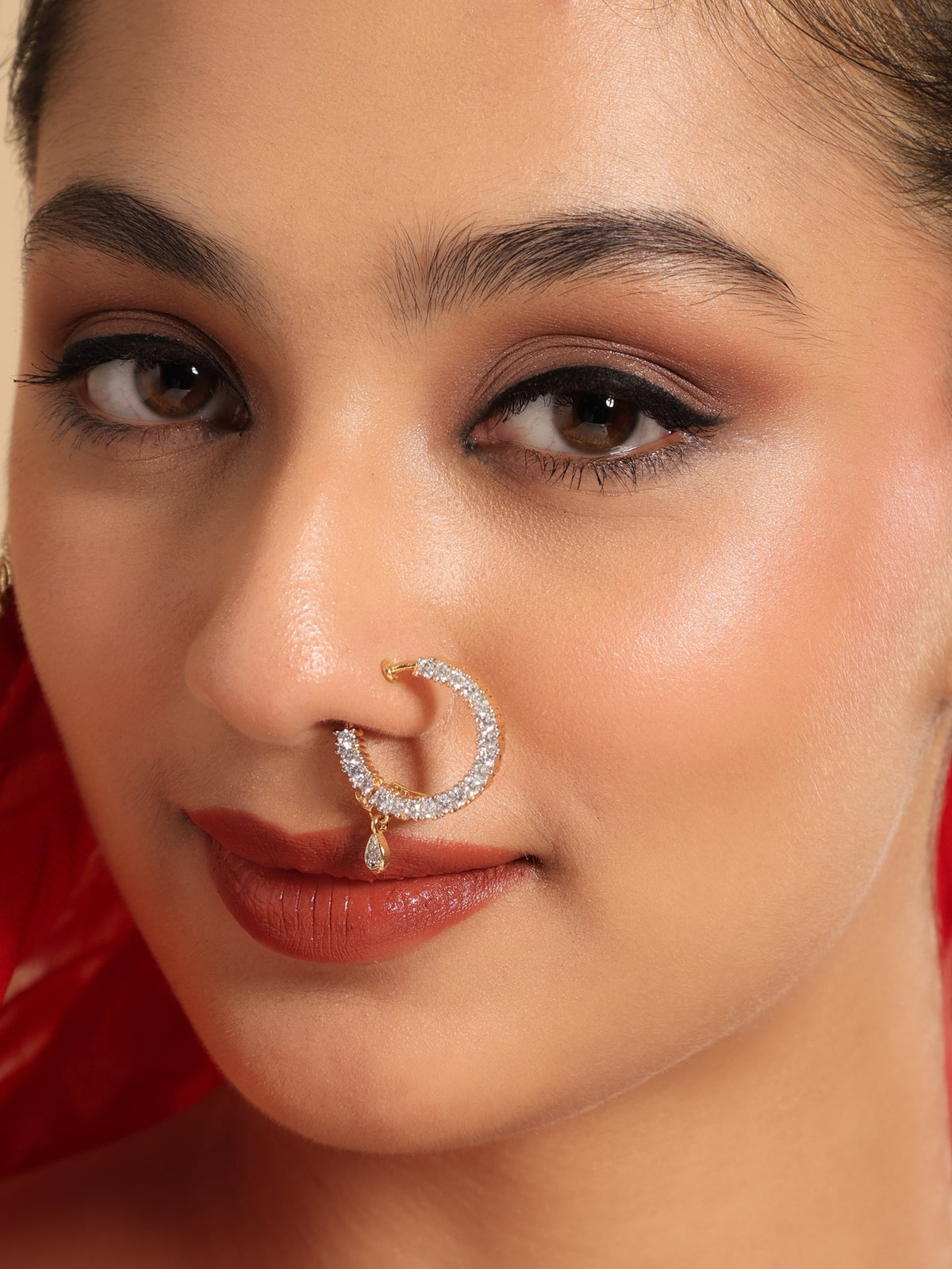 A close-up of a woman wearing a gold crescent-shaped nose ring adorned with small clear stones and a dangling teardrop charm, set against a warm skin tone with a red fabric background.