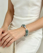 A close-up of a woman wearing a white sleeveless dress, showcasing a beaded wrap bracelet with green and white beads. She is holding a pearl-embellished clutch purse.