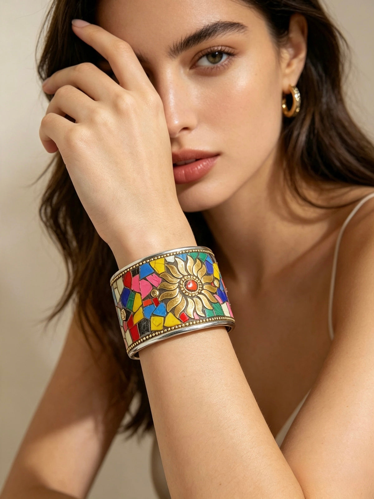 A close-up of a woman wearing a wide silver cuff bracelet decorated with a colourful mosaic floral pattern, set against a warm beige background.