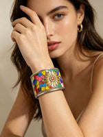 A close-up of a woman wearing a wide silver cuff bracelet decorated with a colourful mosaic floral pattern, set against a warm beige background.
