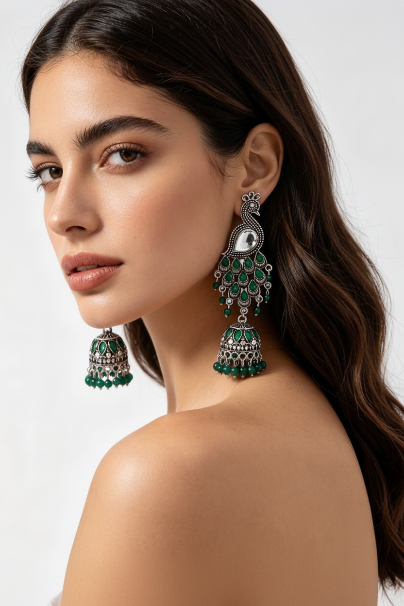 A close-up of a woman wearing a pair of ornate silver-toned dangle earrings with green stone accents and a central white stone, shown against a plain light background.