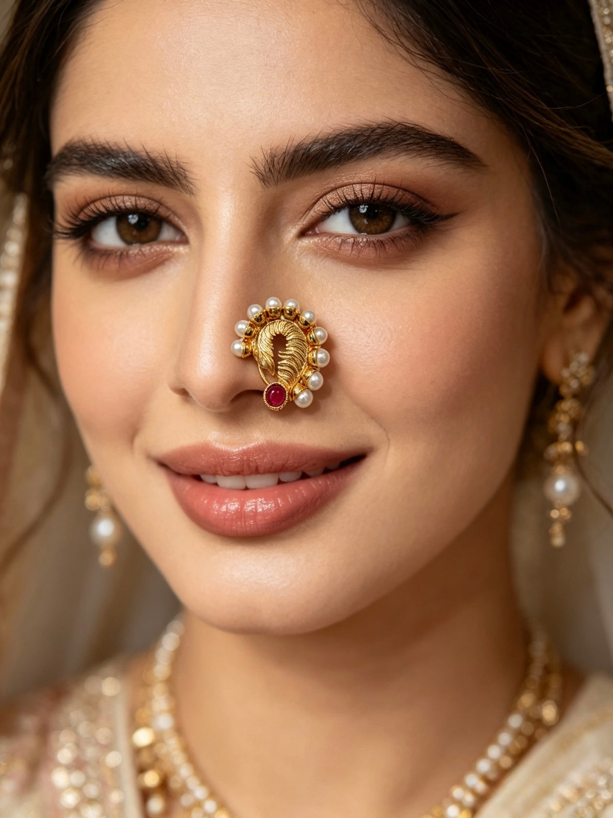 A close-up portrait of a woman wearing traditional Indian jewelry, featuring a gold-plated nose ring (Hastapurna) adorned with white pearls and a small red ruby stone, along with matching pearl and gold earrings.