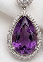 A close-up view of luxury silver-set earrings, showcasing a deep purple pear-shaped amethyst center stone framed by a brilliant pavé diamond halo against a soft white background.