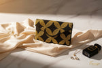 A clutch bag with a geometric pattern of gold and black stars on a dark background, placed on a wooden surface, with a few green leaves scattered around.