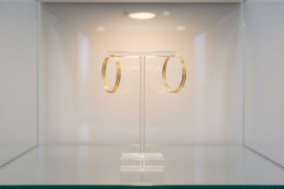 A crisp, professional studio photograph of the 10ct gold-plated baali earrings displayed on a luxury jewellery T-bar stand, showcasing the high-polish finish and the uniform curvature of the brass base.