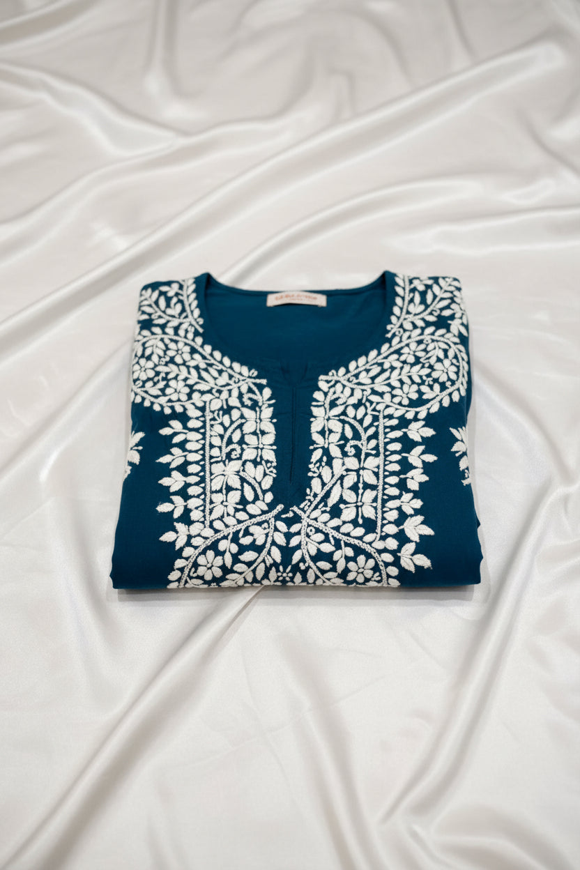A deep dark teal colour code is #215D6E tunic dress with intricate white floral and geometric block-print embroidery, laid flat on a grey wood-look floor.