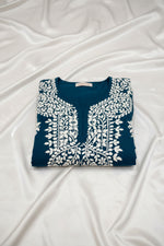 A deep dark teal colour code is #215D6E tunic dress with intricate white floral and geometric block-print embroidery, laid flat on a grey wood-look floor.