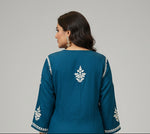 A deep dark teal colour code is #215D6E tunic dress with intricate white floral and geometric block-print embroidery, laid flat on a grey wood-look floor.