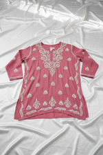 A dusty rose pink long-sleeve tunic top with white floral block prints, hanging on a wooden hanger against a white marble background.