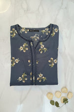 A folded slate blue kurta featuring a cream and green floral print, with a decorative lace trim along the neckline and chest. A black brand label reading Famaza Couture is visible on the inside collar.
