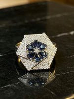 A geometric hexagonal ring featuring a central cluster of deep blue teardrop-shaped gemstones surrounded by a halo of small clear stones, set on a polished silver-toned band and displayed on a dark marble surface.