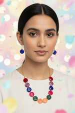 A girl wearing the Facet Octagon Resin Necklace, showcasing the vibrant jewel-toned beads in a mirrored peach, emerald, and magenta pattern. She is modeling the matching cobalt-blue octagon earrings, which dangle elegantly to highlight the set's bold geometric design and high-shine marbled finish.